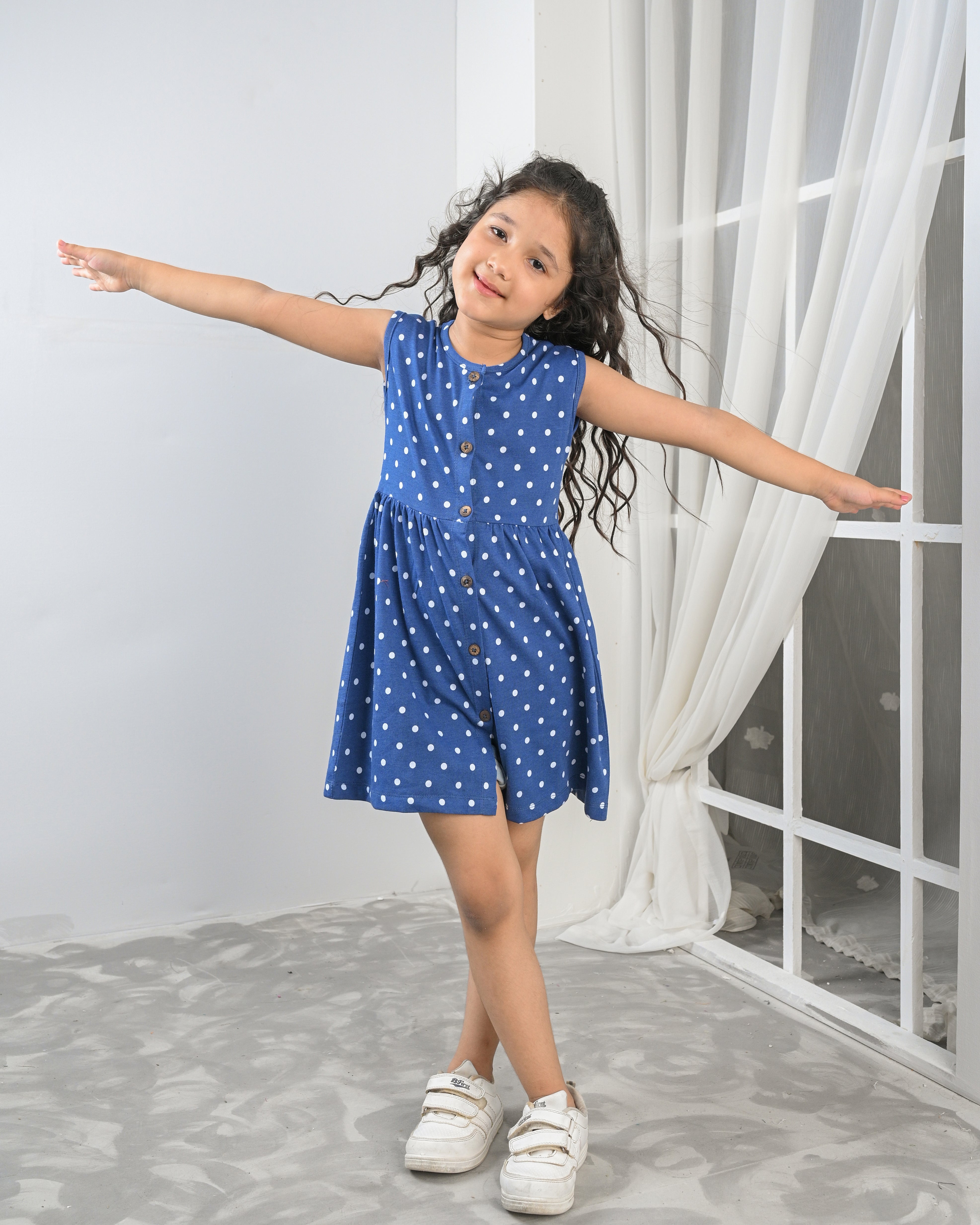 Young girl in a blue polka dot dress standing with arms outstretched in a room with white curtains.